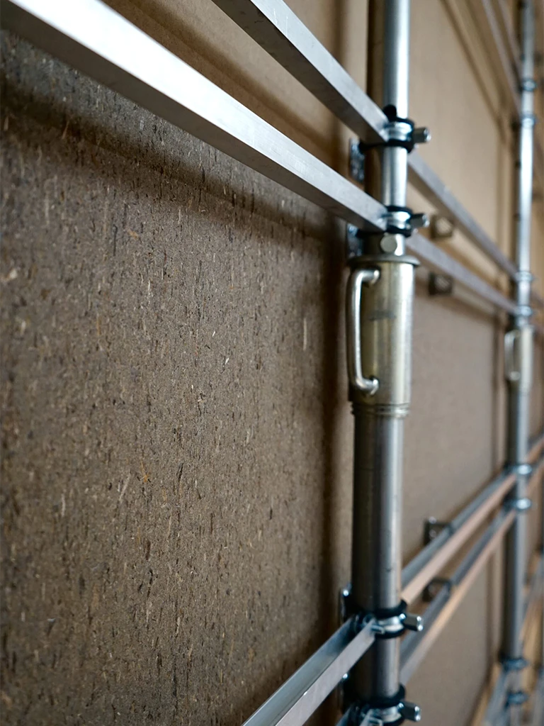 Macro shot showing the metal scaffolding and connector joints of a Mellow Wall installation, mounted against a cork-textured surface.
