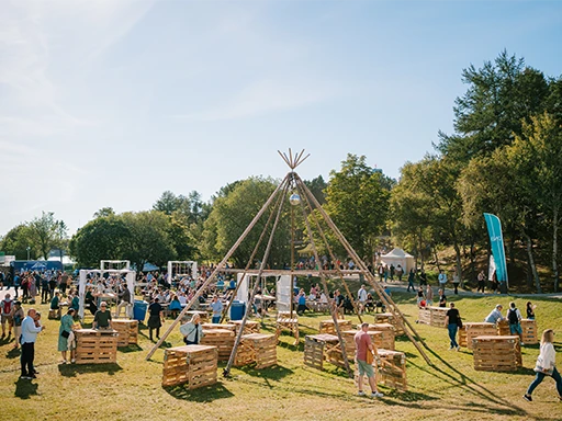 People gathering outdoors around a large wooden tipi structure at a cultural festival set in a grassy park with trees and sunny weather.