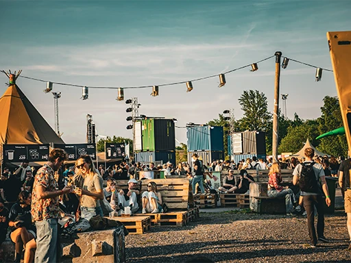 Festivalgoers socializing at an outdoor event with pallet seating, string lights, and colorful shipping container structures in the background.