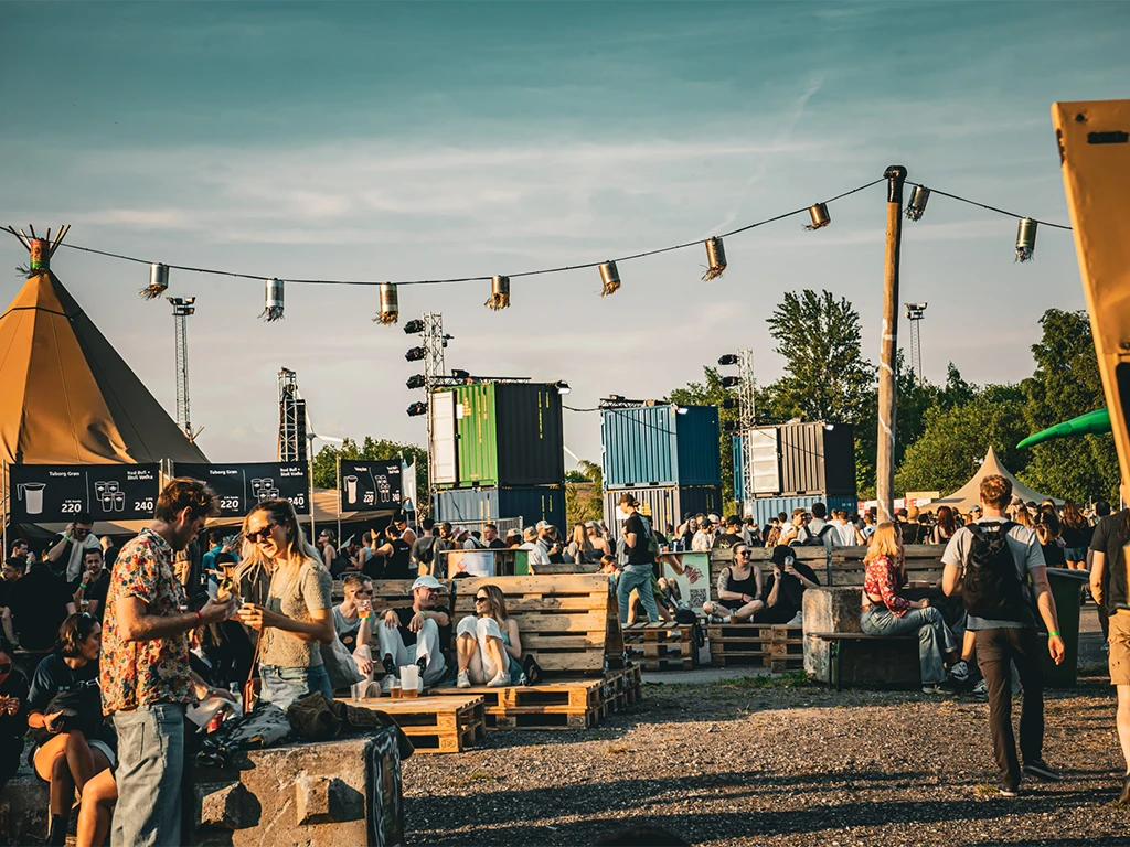 Crowds gather and relax on pallet-based seating at a sunlit outdoor festival, surrounded by colorful shipping containers and string lights.