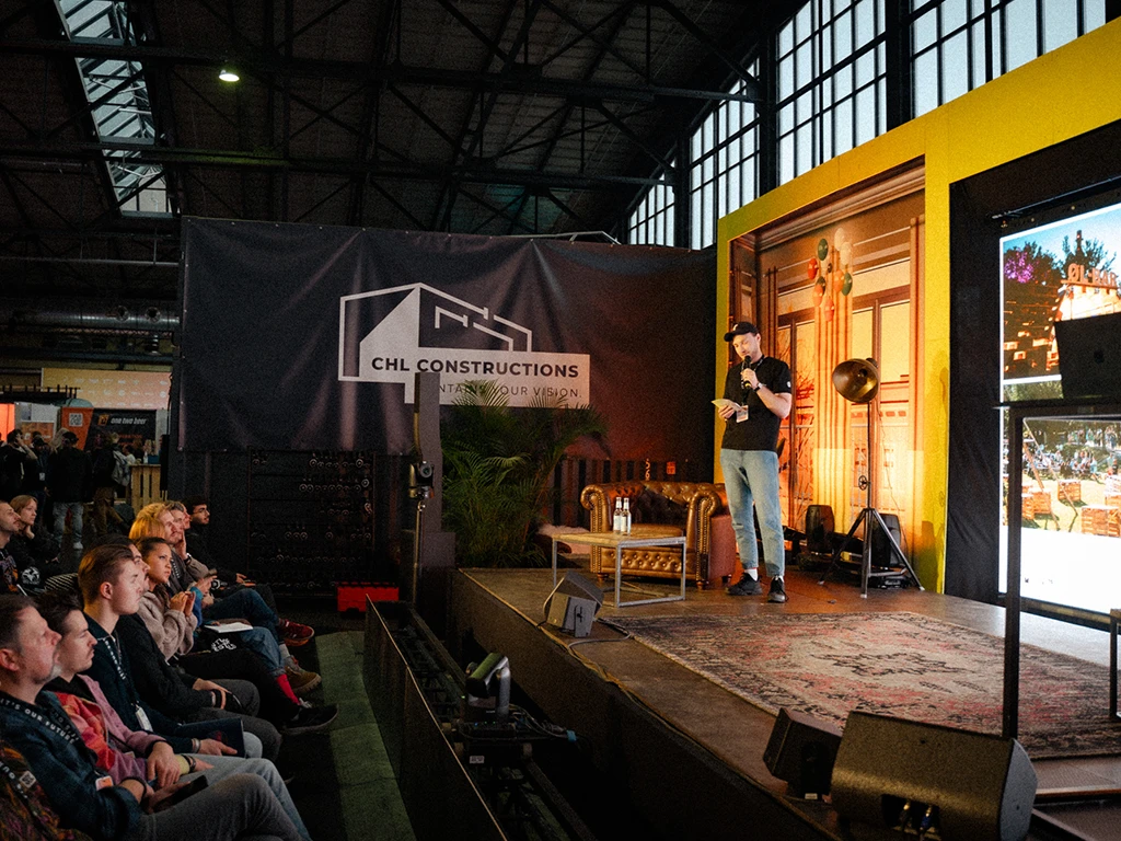 A speaker presents on stage at an indoor event, backed by a branded CHL Constructions banner, with a seated audience attentively listening.