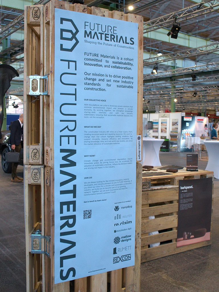 A large vertical banner for 'Future Materials' is mounted on a structure made from upcycled EPAL pallets, surrounded by other exhibition booths in an industrial hall.