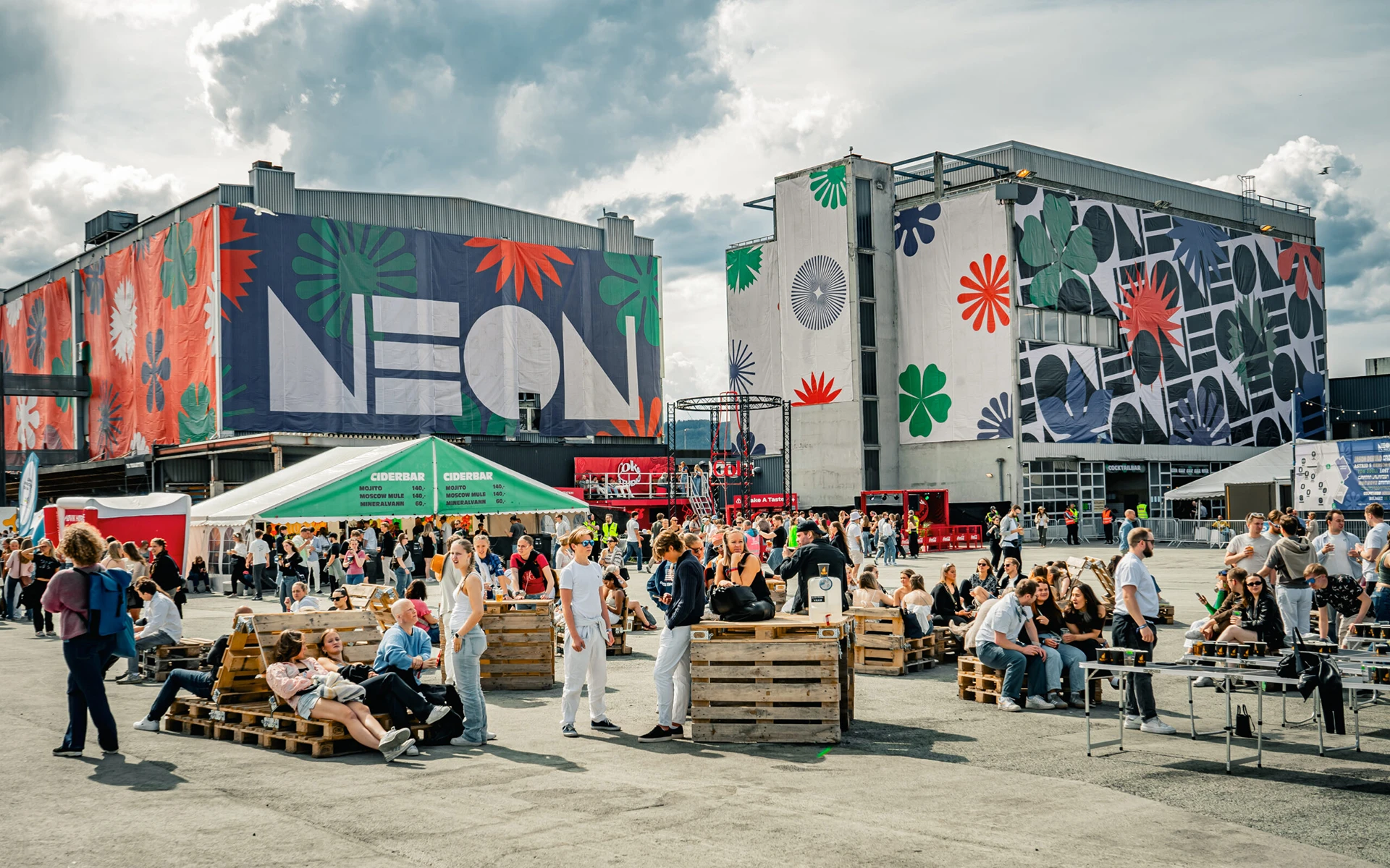 Outdoor festival crowd relaxing on pallet seating in front of large industrial buildings decorated with bold, colorful graphic murals and the word 'NEON' in oversized letters
