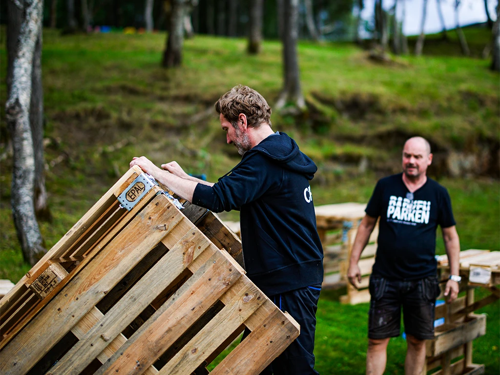 Two men construct a modular setup using EPAL pallets on a grassy outdoor site, highlighting DIY ease and mobility.