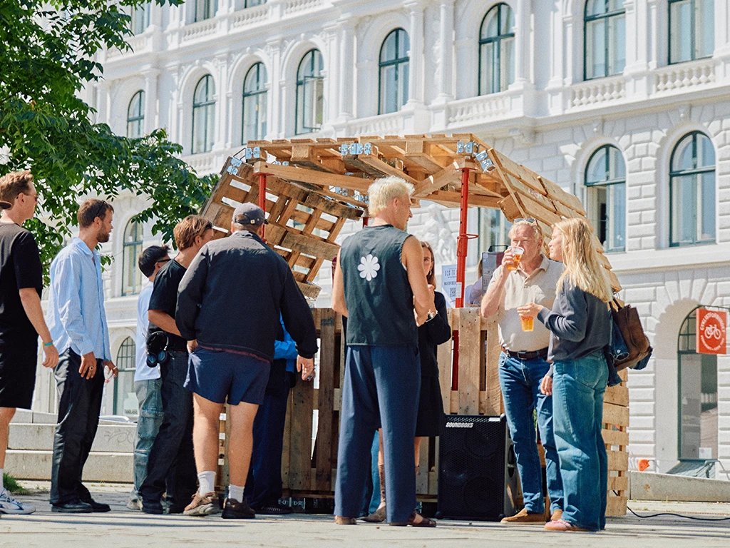 A group of people gathers around a small, arched pavilion structure built from wooden pallets, set in front of a white neoclassical building in a city square.