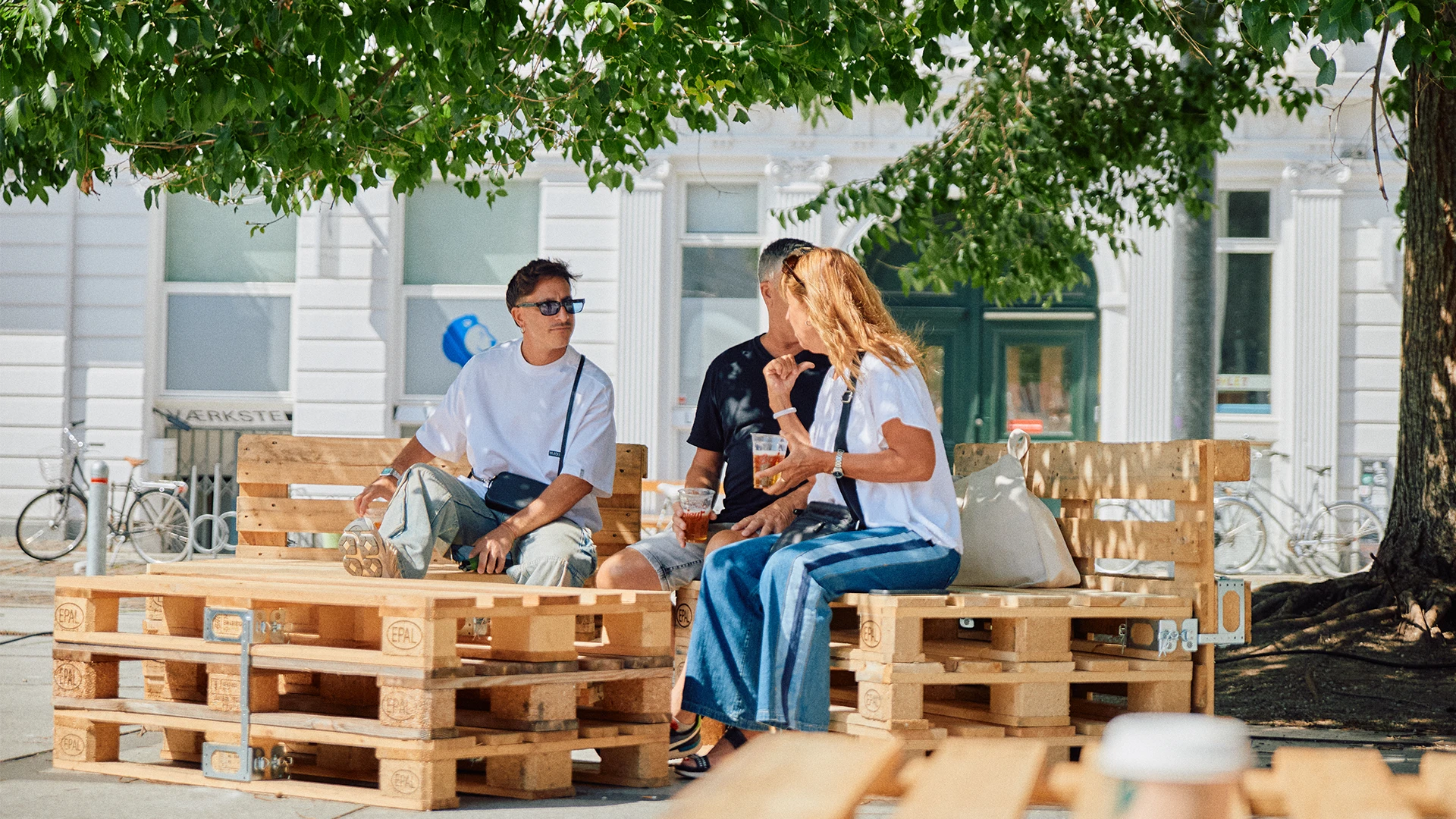 Three people sit and chat on a bench made of stacked pallets in a shaded public space, with bikes and a white building in the background.