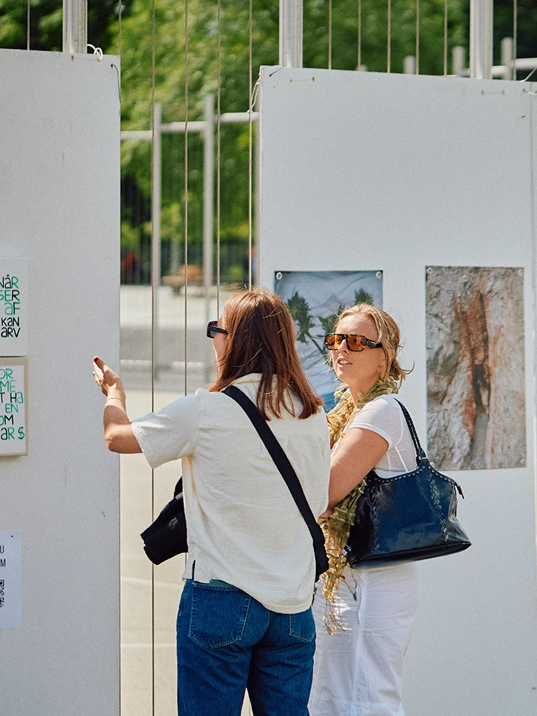 Two women view and discuss posters and photographs mounted on temporary white exhibition panels outdoors.