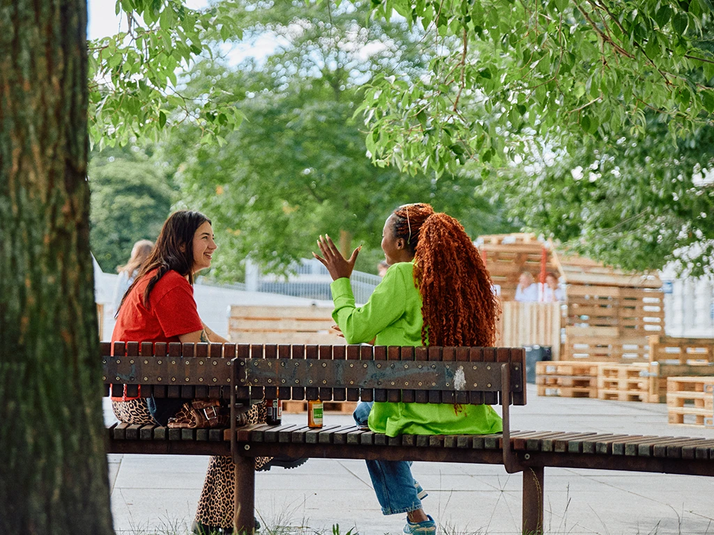 Two women sit on a public bench under leafy trees, talking and laughing, with modular pallet structures and event furniture visible in the background.